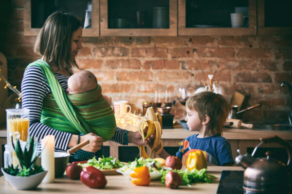 Happy young family, beautiful mother with two children, adorable preschool boy and baby in sling cooking together in a sunny kitchen.